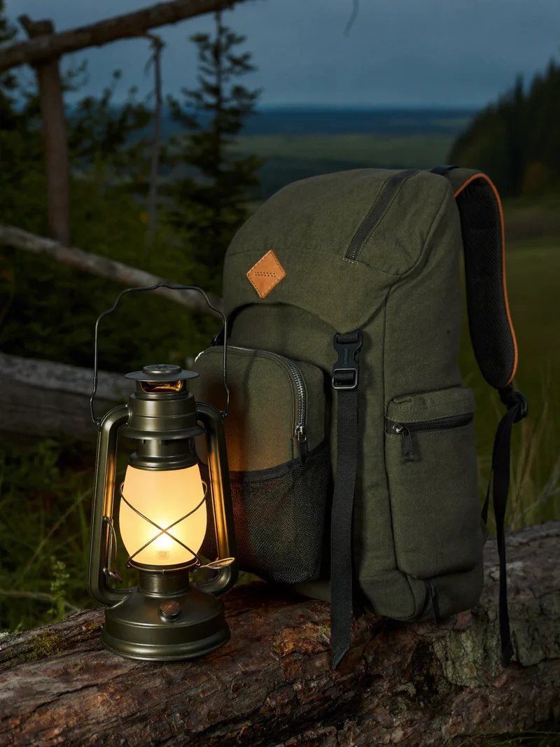 Warme Campinglaterne neben gepacktem Trekkingrucksack auf einem Baumstamm in der Daemmerung